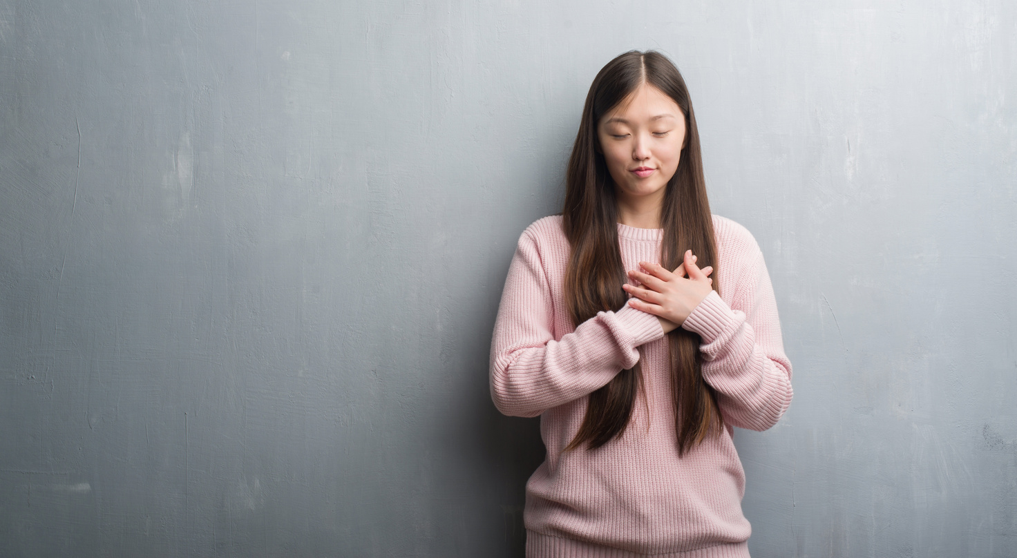 Young Chinese woman over grey wall smiling with hands on chest with closed eyes and grateful gesture on face. Health concept.